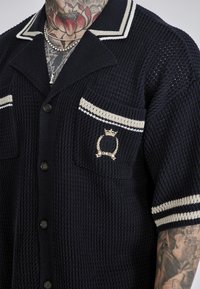 Maglietta blu marina a maglia con vestibilità rilassata, caratterizzata da un colletto a punte, due tasche sul petto con strisce crema a contrasto e dettaglio del logo ricamato.