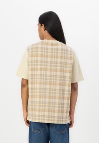 Kortermet skjorte med et beige rutemønster på baksiden og en ensfarget lys beige front. Stoffet er glatt og har en avslappet passform.