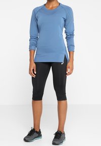 Niebieska, długorękawowa koszulka sportowa z okrągłym dekoltem, zestawiona z czarnymi legginsami do kolan i czarnymi butami do biegania z turkusowymi akcentami.