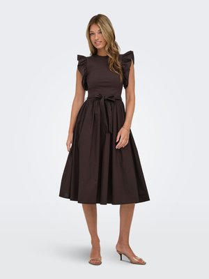 Femme portant une robe midi noire sans manches avec des épaules volantées, une ceinture à la taille et des sandales à talons beiges ouvertes à l'avant, debout sur un fond uni.
