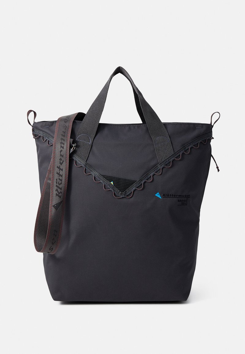 Black tote bag made of textured fabric; features dual handles, a decorative scalloped edge, and an adjustable shoulder strap. Logo displayed.