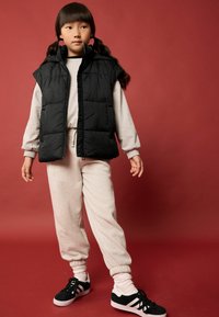 Svart pufferväst med hög krage, grå sweatshirt, beige sweatpants och svarta sneakers med rosa ränder. Bakgrunden är röd.