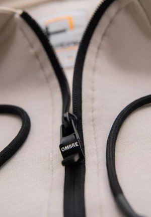 Beżowy materiał z czarnym suwakiem i sznurkiem, z uchwytem do suwaka oznaczonym napisem "OMBRE". Gładka struktura i widoczne detale czystego przeszycia.