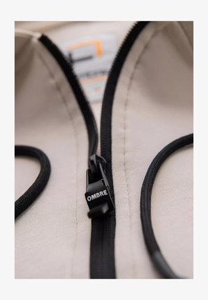 Beżowy materiał z czarnym suwakiem i sznurkiem, z uchwytem do suwaka oznaczonym napisem "OMBRE". Gładka struktura i widoczne detale czystego przeszycia.