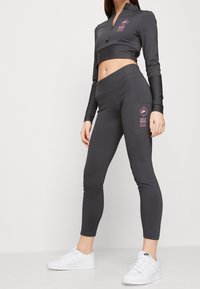 Kvinna klädd i mörkgrå, kort zip-up topp och matchande leggings med rosa logotyper, tillsammans med vita Nike-sneakers, som står mot vit bakgrund.