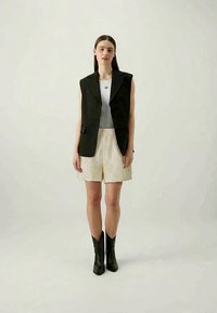 Musta hihaton bleiseri, valkoinen ribattu toppi, kermaiset tekstuurishortsit ja mustat nilkkasaappaat. Yksinkertainen, moderni muotoilu, jossa on rakenteellinen istuvuus.