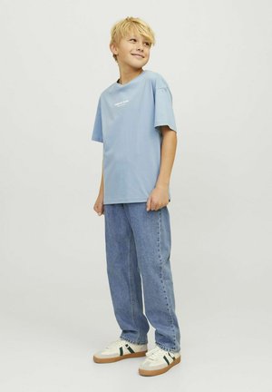 Lichtblauwe katoenen t-shirt met korte mouwen en witte tekst, gecombineerd met losse blauwe jeans en witte sneakers met groene accenten.