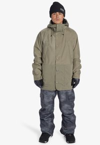 Khaki vandtæt jakke med hætte, trykknapper og for- lommme; parret med mørkegrå mønstrede skibukser og sorte støvler.