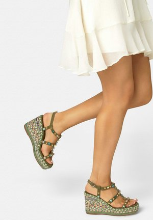 Piernas con falda blanca con volantes y sandalias cuña verdes con joyas, suelas plataforma trenzadas y multicolores.