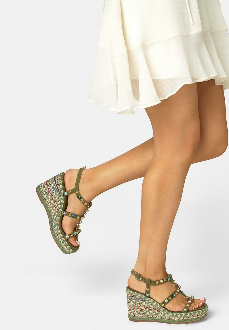 Jambes portant une jupe blanche à volants et des sandales compensées vertes ornées de bijoux, avec des semelles compensées tressées multicolores.