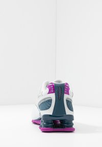 Bílé sportovní boty s šedými a fialovými akcenty, s texturovaným svrškem, polstrovaným límcem a pružným, zřetelným designem podpatku.