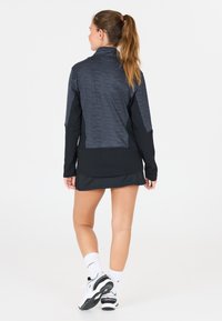 Kvinna med långt hästsvans iförd en svart och grå jacka, svart kjol, vita strumpor och vita sneakers, stående vänd bort på en vit bakgrund.