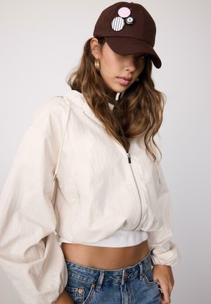 Jonge vrouw met een bruine pet met pins, gouden oorringen, een witte cropped jas met ritssluiting en blauwe spijkerbroek tegen een effen achtergrond.