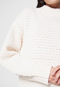 Pull tricoté couleur crème avec un motif texturé, col rond côtelé et manches longues. Design doux à tissage ouvert.