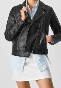 Giacca da biker in pelle nera con accenti trapuntati, zip argentate e un design aderente. Indossata sopra una camicia a righe blu chiaro e una gonna bianca.