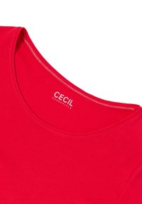 Röd kortärmad topp med rundad halsringning. Tillverkad av mjukt tyg med slät textur. Märkesnamnet "CECIL" tryckt i vitt på kragen.