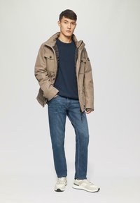 Jasnobrązowa kurtka z kapturem, niebieski sweter oraz niebieskie dżinsy. Wyróżniają się przednimi kieszeniami oraz beżowymi butami sportowymi z szarymi i zielonymi akcentami.