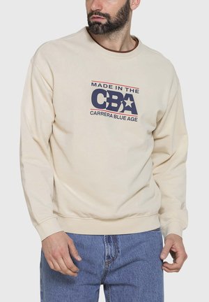 Uomo con barba che indossa una felpa beige con la scritta "Made in the CBA Carrera Blue Age" e jeans blu, guarda di lato.