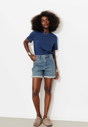 Femme appuyée contre un mur blanc, portant un t-shirt bleu, un short en jean retroussé, des chaussures beige slip-on, des bracelets manchette en or et un collier chaîne en or.