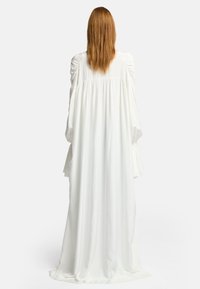Langes, fließendes weißes Kleid mit langen Ärmeln, gerafften Schulterdetails und einer weichen Textur, das in einer entspannten Silhouette bis zum Boden fällt.