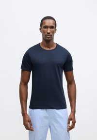 Temno modra majica s kratkimi rokavi iz gladkega blaga z okroglim izrezom in majhno logotipno podrobnostjo spredaj, kombinirana s svetlimi karo kratkimi hlačami.