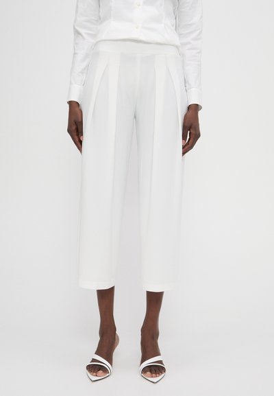 Weiße Hosen mit hoher Taille, gepunktetem Design und verkürzter Länge. Der Stoff ist glatt und hat ein sauberes Finish. Kombiniert mit weißen Absatzsandalen.