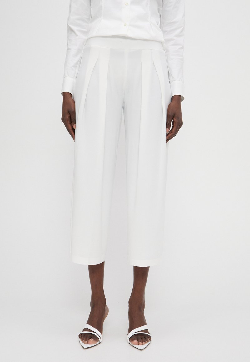 Weiße Hosen mit hoher Taille, gepunktetem Design und verkürzter Länge. Der Stoff ist glatt und hat ein sauberes Finish. Kombiniert mit weißen Absatzsandalen.