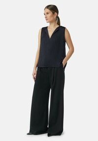 Top senza maniche a pieghe di colore blu navy con scollatura a V e tasche laterali, abbinato a pantaloni neri a gamba larga e scarpe con tacco.