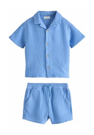 Tout-petit portant une chemise à boutons texturée bleu clair à manches courtes et un short assorti, avec des sandales à boucle marron sur un fond clair.