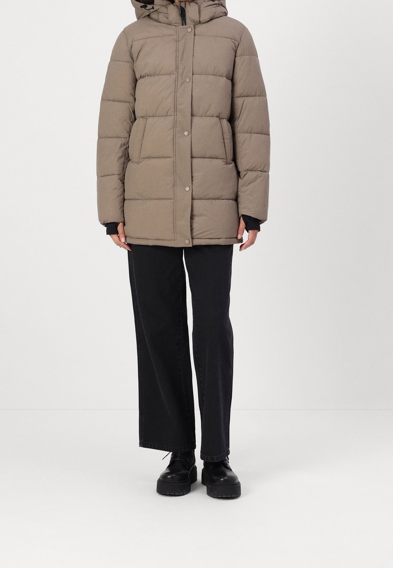 Personne portant une veste rembourrée marron à poches avant, un pantalon noir à jambes larges et des bottes noires épaisses à lacets, debout devant un fond uni.