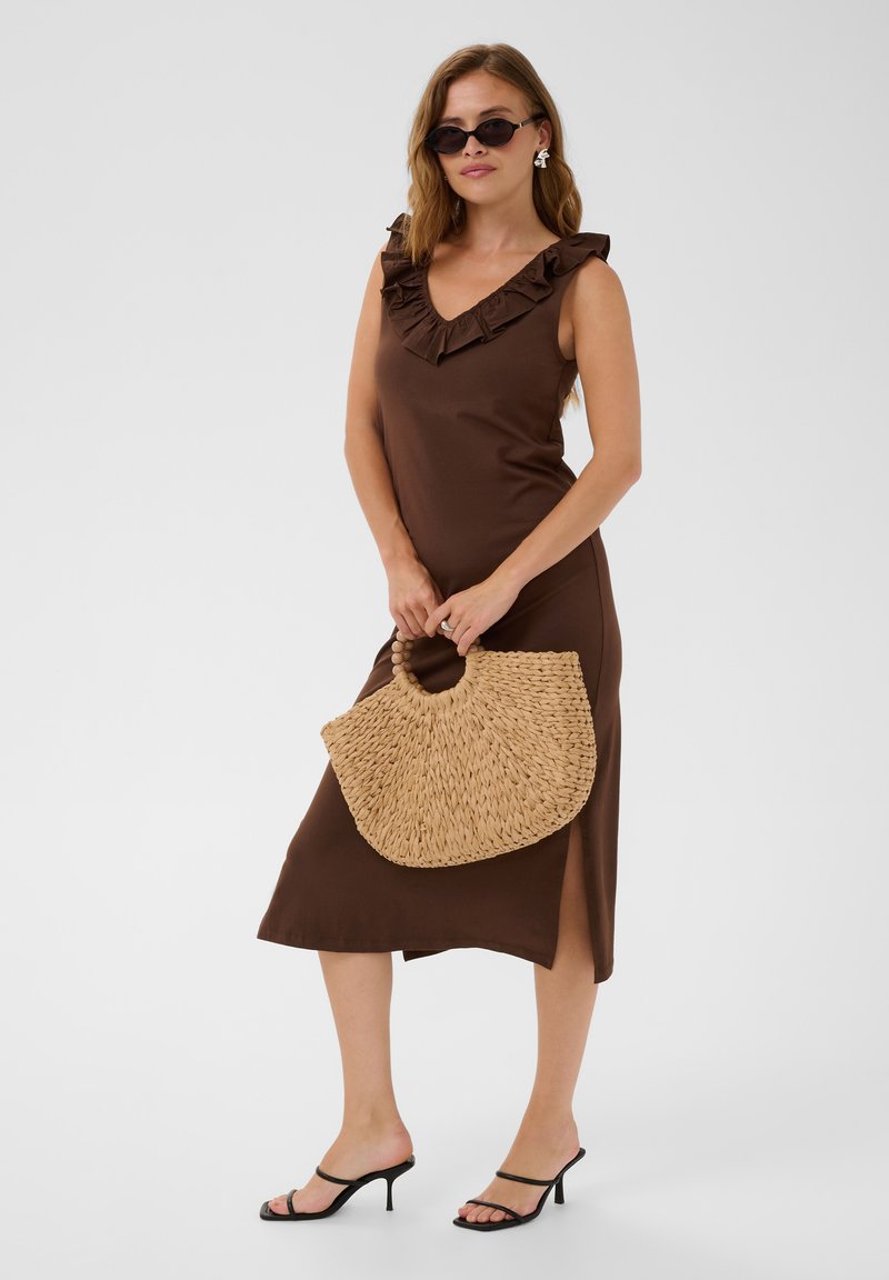 Femme portant une robe marron sans manches avec un col volanté, des sandales noires à talons, des lunettes de soleil, des boucles d'oreilles et tenant un sac à main en paille tressée.