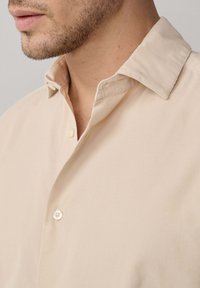 Chemise beige à boutons avec un tissu texturé, dotée d'un col replié et d'un bouton visible près de l'encolure.