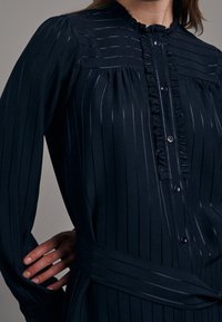 Mörkblå knäppt klänning med vertikala ränder, volangkrage och matchande bälte. Tillverkad av lätt tyg, med långa ärmar och manschetter.