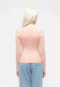 Γυναίκα με μακριά ξανθά μαλλιά, που φοράει ένα εφαρμοστό ροζ παλιό ροζ μακρυμάνικο τοπ και ανοιχτό μπλε υφασμάτινο παντελόνι, γυρισμένη προς τα πίσω σε λευκό απλό φόντο.