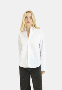 Camicia bianca con colletto a punta, caratterizzata da sottili punti metallici sparsi. Maniche lunghe con polsini abbottonati, indossata con pantaloni scuri.