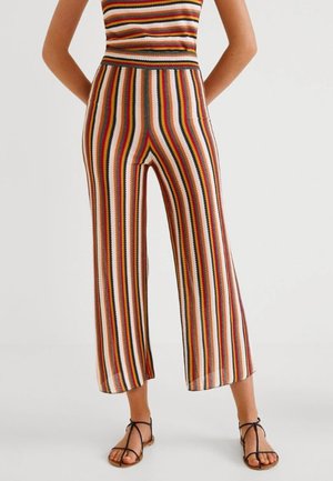 Femme portant un pantalon en maille à rayures verticales multicolores avec un haut sans manches assorti et des sandales plates noires à lanières, debout devant un fond blanc.