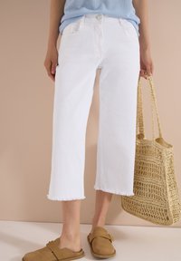 Mujer con pantalones blancos cortos con dobladillo deshilachado, camiseta azul clara, zapatos sin cordones color beige y llevando un gran bolso de paja tejido.