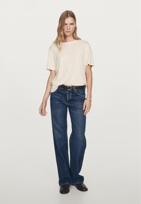 Beige tričko s krátkým rukávem v kombinaci s tmavě modrými širokými džínami a hnědým opaskem. Modelka stojí sebevědomě na neutrálním pozadí.