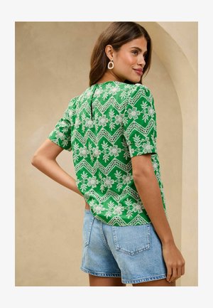 Zelená tričko s krátkym rukávom s bielymi kvetinovými a cik-cak vzormi, s detailom kľúčovej dierky na chrbte. Skombinované s modrými džínsovými šortkami.
