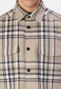 Beige plaid shirt made of textured fabric, featuring a classic collar, dark buttons, and chest pockets with a contrast check pattern.