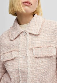 Vaaleanpunainen tweed-takki, jossa on tekstuuri, kauluksessa, neljä etutaskua ja nappikiinnitys. Hapsuttuneet reunat lisäävät yksityiskohtia.