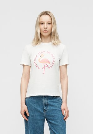 Ξανθιά γυναίκα που φοράει λευκό μπλουζάκι με ροζ φλαμίνγκο και κείμενο «Άλλη μια μέρα στον παράδεισο», συνδυασμένο με μπλε τζιν, στέκεται μπροστά σε απλό φόντο.