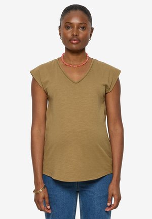 Frau steht frontal, trägt ein olivgrünes V-Ausschnitt-Oberteil mit kurzen Ärmeln, blaue Jeans, eine rosa Perlenkette und goldene Armreifen.