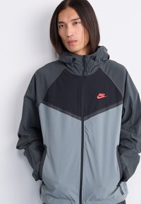 Nike windbreaker jacket featuring a color-block design in gray and black. It has a high collar, zip closure, and two side pockets.