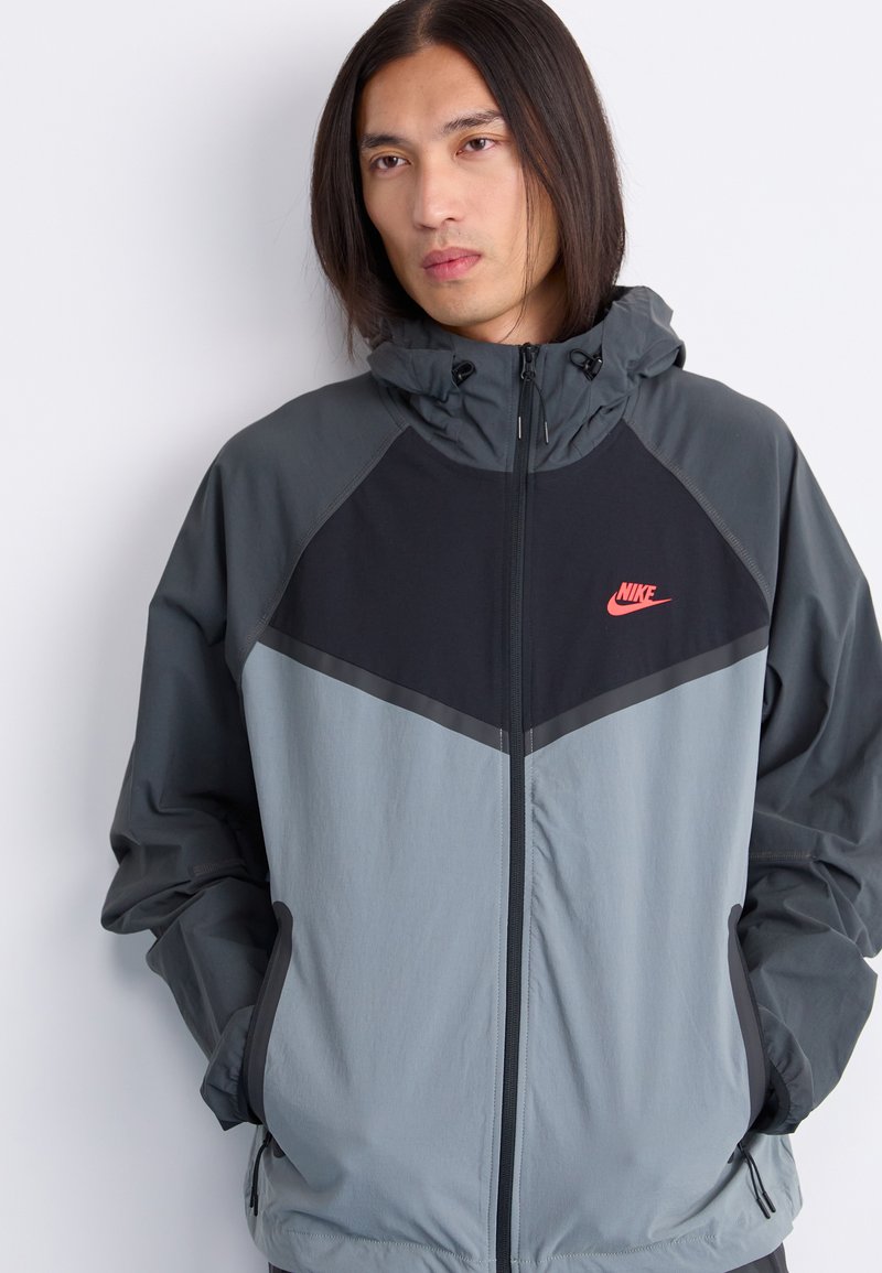 Nike kurtka przeciwwiatrowa z kolorowym blokiem w odcieniach szaro-czarnych. Posiada wysoki kołnierz, zamek błyskawiczny oraz dwie boczne kieszenie.