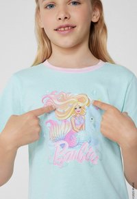 Svetle modré triko s krátkým rukávem a růžovým límečkem, s barevnou grafikou mořské panny a textem "Barbie" v růžové, ukazující prsty na design.