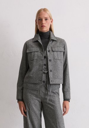 Veste grise raccourcie avec un col, deux poches poitrine et fermeture à bouton, fabriquée en tissu mélangé en laine texturé. Portée par-dessus un col roulé gris.