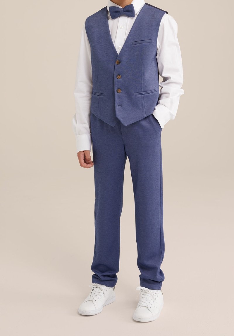 Blå väst med tre knappar och fickor, vit skjorta, matchande blå byxor och vita sneakers. Slät textur, skräddarsydd passform och klassisk design.