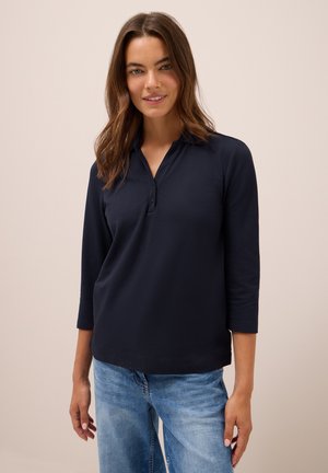 Marineblauw langarm polo shirt van zachte stof, met een knoopsluiting op de kraag en een relaxte pasvorm. Gecombineerd met lichtblauwe denim jeans.