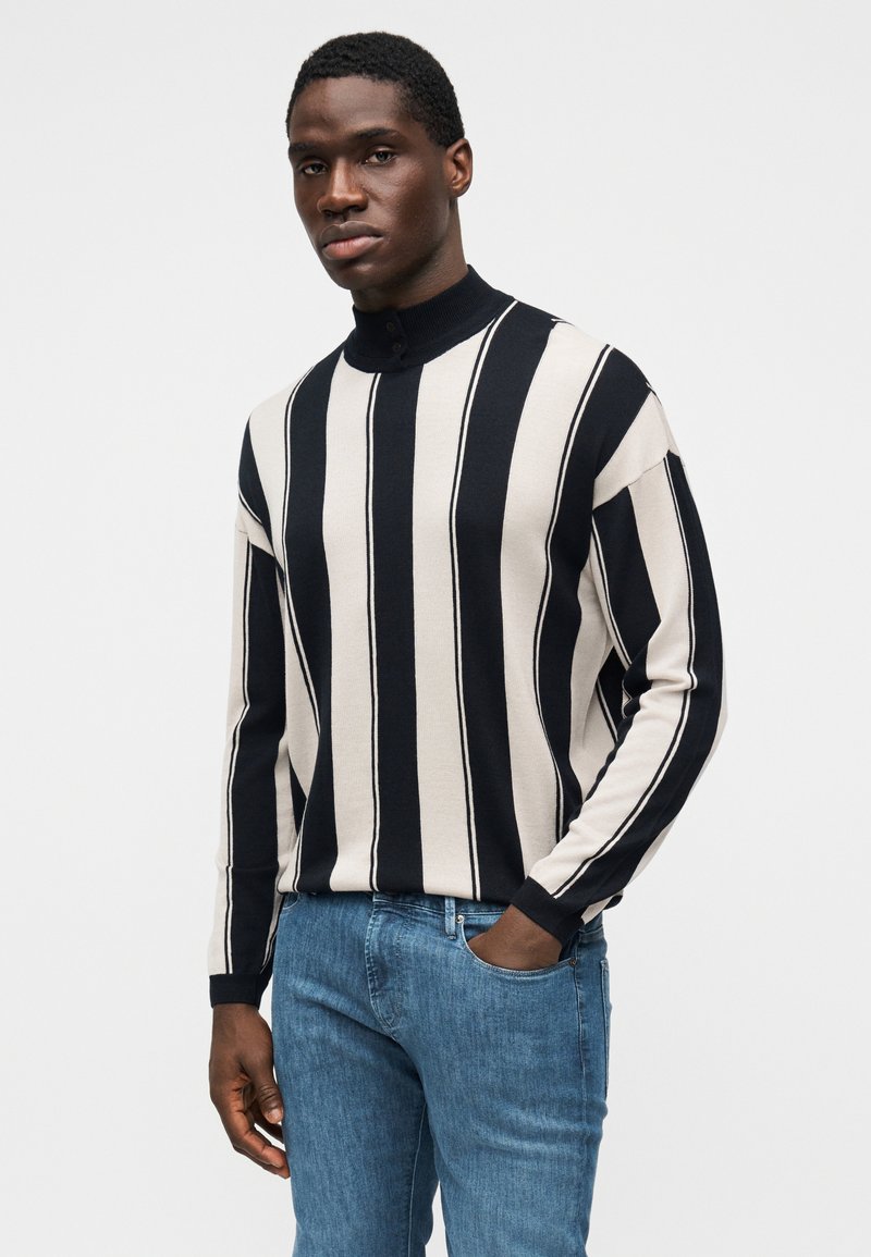 Pull rayé noir et beige à col montant et manches longues, associé à un jean en denim bleu. Texture douce et design structuré.